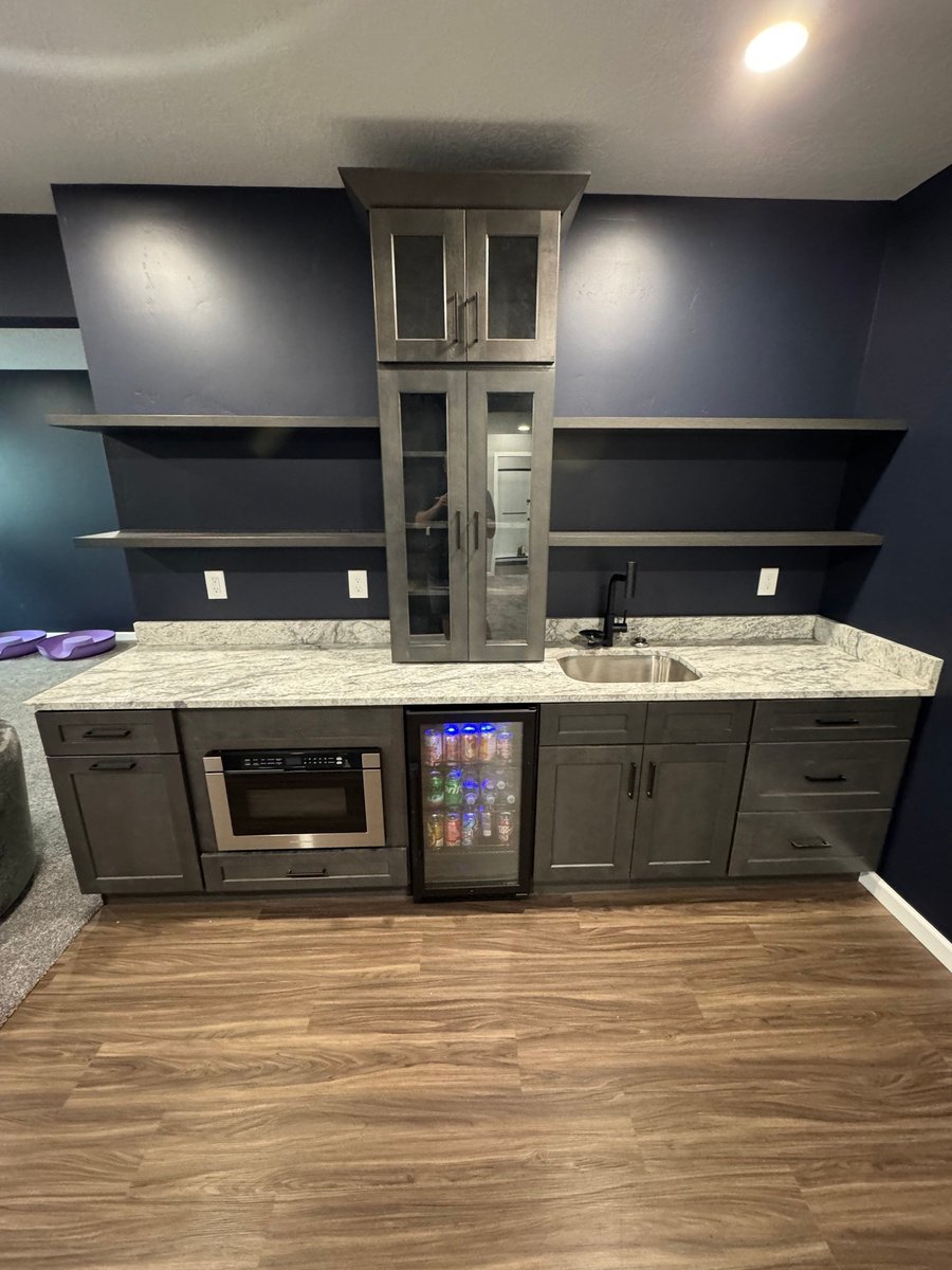 Finished custom basement wet bar with dark cabinetry, stone countertop, sink, microwave, beverage fridge, and open shelving.