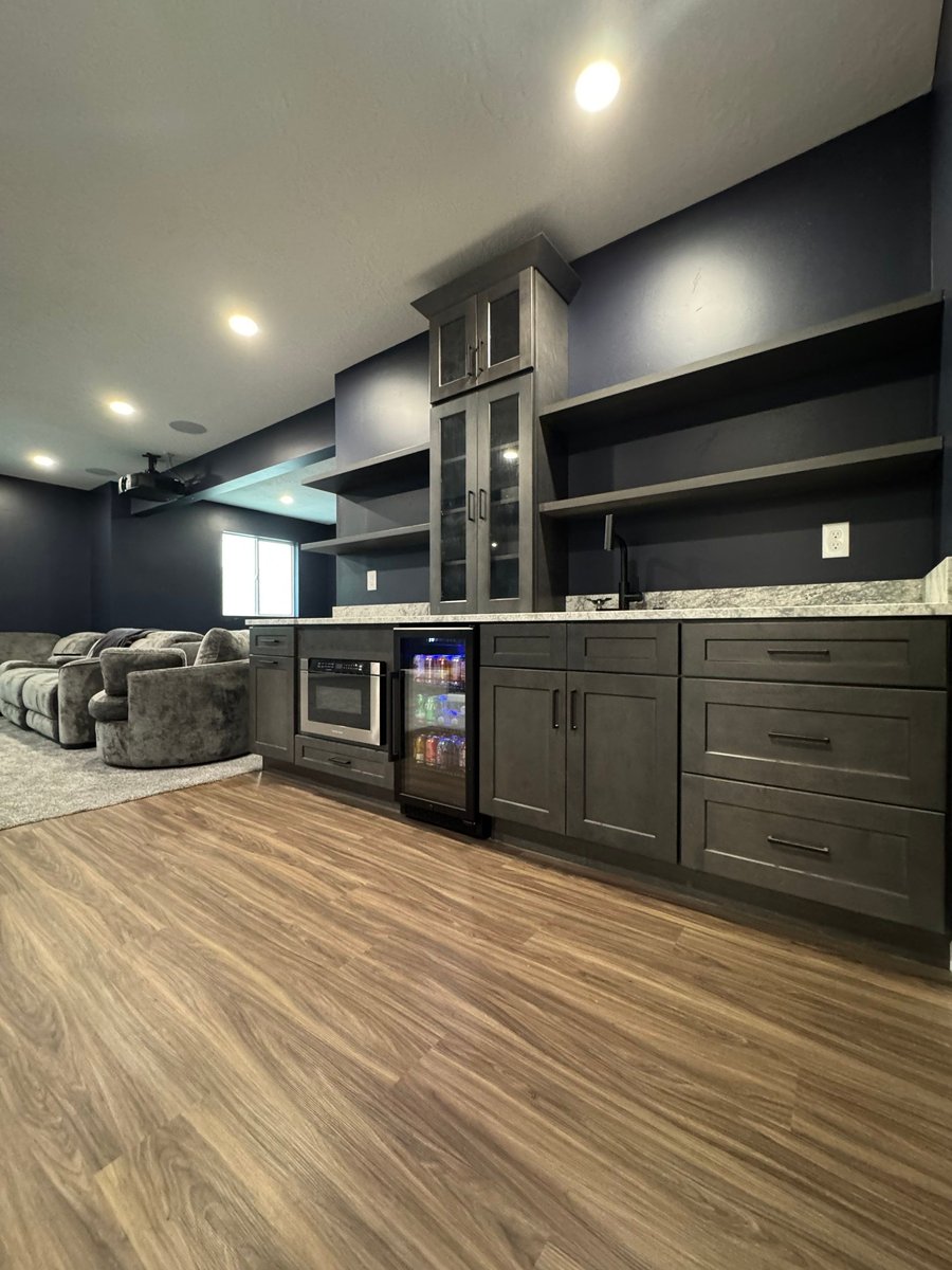 Custom basement wet bar with dark cabinetry, beverage fridge, sink, and shelving.